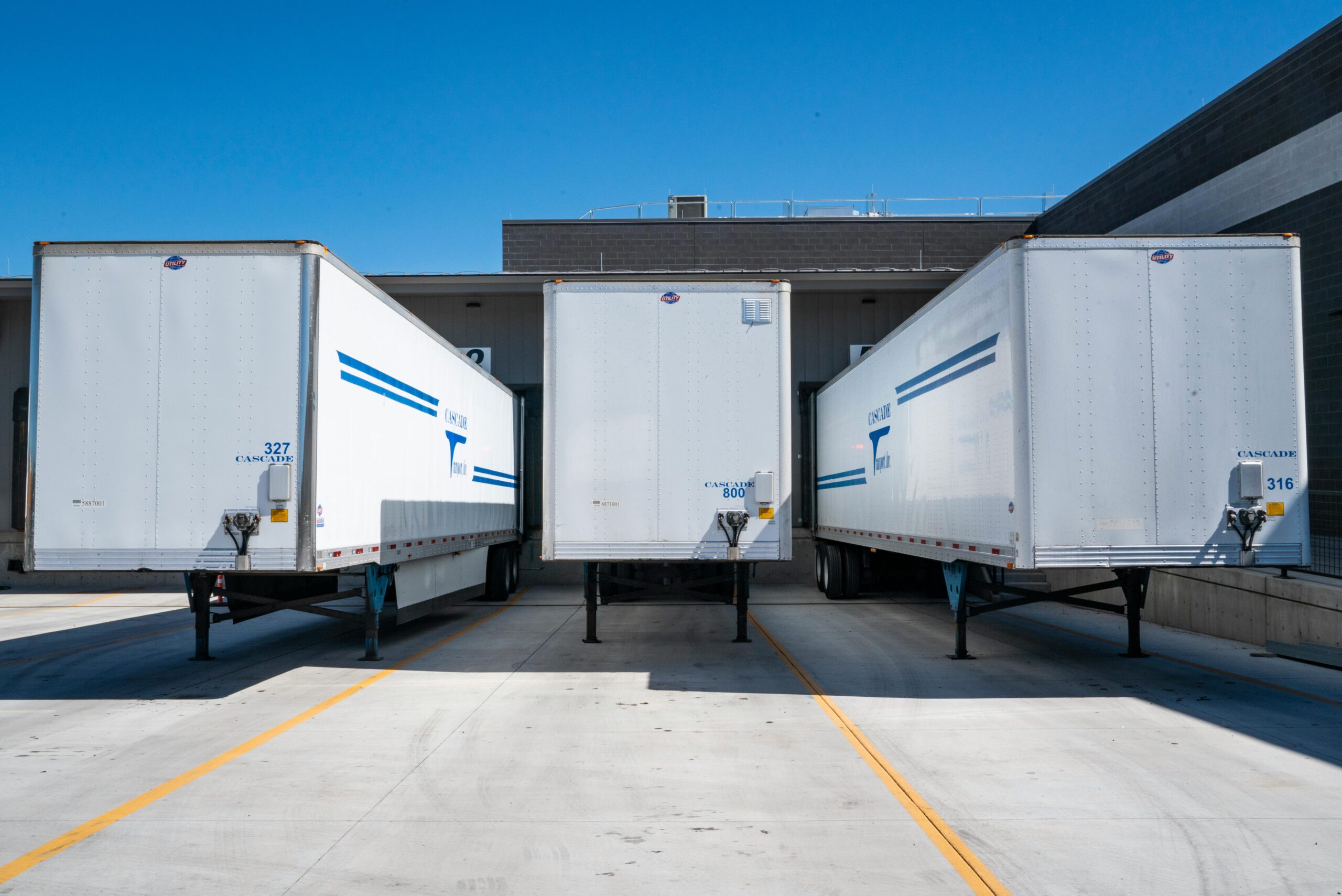 Home Three white cargo trailers parked at an industrial shipping dock under clear blue skies.